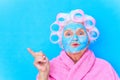 old woman wirh pink curlers on head and blue hyaluronic beauty mask on face wear cotton robe, preparing for a date