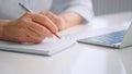 Old woman wrinkly hands write in paper notebook with pen at table near grey laptop keyboard