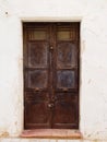 Old wooden brown double doors with chipped flaking faded peeling paint and rusty handles padlocked closed in a white painted wall