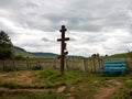 Old wooden cross stands sideways near a crooked fence and a bench against the backdrop of a village on a cloudy summer.