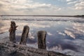 Old wooden posts left after mining salt on the shore of a salt lake. Reflection of clouds in the water surface.