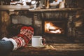 Old wooden retro table top with woman`s legs and a mug and a book and fireplace background.