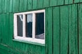 An old wooden small window with a frame of boards painted white and thin glass. On a battered wall of wooden planks with green pai Royalty Free Stock Photo