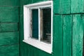 An old wooden small window with a frame of boards painted white and thin glass. On a battered wall of wooden planks with green pai Royalty Free Stock Photo