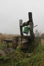 An old wooden well with a green bucket. Foggy rural landscape against the backdrop of an autumn field and village