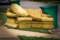 Old yellow couch with cushions sits on a curb in residential area next to a recycling cart waiting for bulk waste hauler pickup on