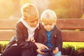 Older brother teaches younger to use the mobile phone. Kids sitting on bench. Happy children playing games on smartphone outdoors Royalty Free Stock Photo