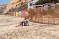 Older couple baby boomers sit on beach under umbrella near a chain link fence with dilapidated houses in the background Royalty Free Stock Photo