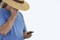 Older long gray haired man in wide brim straw hat and tee shirt shade his eyes to see cell phone - isolated on white