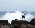 Older man fishing in the sea stood on rocks with dramtic waves a