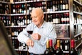 Older man inspecting quality of red wine in wine store in search of perfect wine for solemn occasion