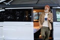 Older man traveler standing near rv camper van drinking coffee enjoying view.