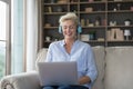 Older pretty woman wear headphone sit on sofa with laptop