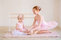 The older sister, a ballerina in a pink tutu, puts pointe shoes on the baby while sitting at the barre