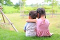 Older sister hugs little brother by the neck, shoulders sitting on green grass field. Two adorable Asian children sitting and