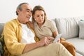 Older spouses using cellphone, browsing internet or video calling with family, sitting on couch at home, free space