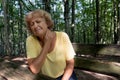 An older woman expresses neck pain. Sad mature blonde woman sitting on a bench in the forest after walking and suffering from neck Royalty Free Stock Photo