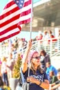 Older woman with flag and Womens March tee shirt at political rally with crowd blurred behind her