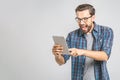 OMG! Great news! Surprised young man in plaid shirt standing and using tablet over grey background