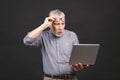 OMG! I`m shocked! Scared senior aged bearded old man in eyeglasses holding laptop computer isolated against black background