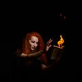 An ominous witch sets her spell book on fire with the power of thought. Red-haired woman conjures for Halloween. Flames Royalty Free Stock Photo