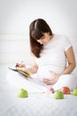 One european pregnant woman writes some idea or thouths in note book by pen sitting on white bed of light room at sunny day. Royalty Free Stock Photo