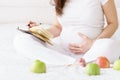 One european pregnant woman writes some idea or thouths in note book by pen sitting on white bed of light room at sunny day. Royalty Free Stock Photo