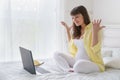 One european pregnant woman with yelow shirt has much emotions from screen of laptop while she is sitting on bed in white bedroom