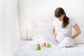 One european pregnant wooman is sitting on white bed with three green apples and smilng touching her abdomen in bedroom Royalty Free Stock Photo