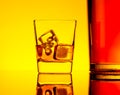 One glass of whiskey with ice cubes near bottle on table with reflection, warm yellow tint atmosphere Royalty Free Stock Photo