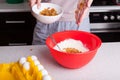 In one hand, girl holds a white cup with raisins in the other grapes themselves and spills them in red bowl with whipped dough,