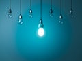 One hanging energy saving light bulb glowing stand out from unlit incandescent bulbs with reflection on dark cyan background Royalty Free Stock Photo