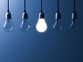 One hanging light bulb glowing different and standing out from unlit incandescent bulbs with reflection on dark blue background Royalty Free Stock Photo