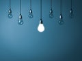 One hanging light bulb glowing and standing out from unlit incandescent bulbs on dark cyan background Royalty Free Stock Photo