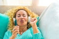 One happy and young woman singing and listening music on the couch or sofa at home with white headphones - music theraphy