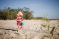 One little girl walks on the beach with a bucket in hand. Barefoot child in panama hat and red dress. Royalty Free Stock Photo