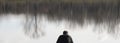 One man in black sitting on a lake bank, with tree reflections on water