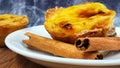 One Pastel de nata or Portuguese egg tart and cinnamon sticks on a white plate. Pastel de Belm is a small pie with a