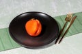 One ugly orange persimmon on black plate and golden cutlery on napkin on light backdrop. Royalty Free Stock Photo