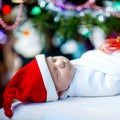 One week old newborn baby in Santa hat wrapped in blanket near Christmas tree with colorful garland lights on background Royalty Free Stock Photo