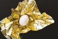 One white egg wrapped in metallic shiny gold paper on black background