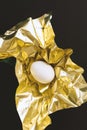 One white egg wrapped in metallic shiny gold paper on black background