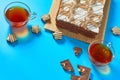 One whole checkered rectangular cake near broken chocolate bar, candy and two full glass cups of tea lies on blue table on party