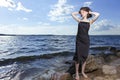 One Winsome Relaxing Caucasian Brunette Girl Posing in Black Dress With Lifted hands On Stone At Sea During Sunny Day Outdoors