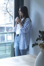 One woman at home standing and looking outside the window drinking cup of coffee and smiling. One female young adult people living Royalty Free Stock Photo