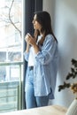 One woman at home standing and looking outside the window drinking cup of coffee and smiling. One female young adult people living Royalty Free Stock Photo