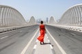 One woman in an orange red dress walking by an empty road on a Meydan bridge with city view on background in Dubai