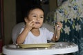 One 1 year old Asian baby boy learning to eat by himself by spoon, messy on baby dining chair Royalty Free Stock Photo