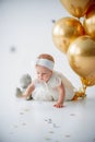 A one year old, baby girl sitting with a bunch of golden balloons and taddy bear on white background in studio Royalty Free Stock Photo