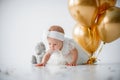 A one year old, baby girl sitting with a bunch of golden balloons and taddy bear on white background in studio Royalty Free Stock Photo